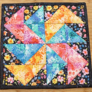 May include: Square quilted hot pad with a colorful geometric design. The center features triangles in shades of pink, blue, orange, and yellow. The border is black with a floral pattern. A decorative item for the kitchen.