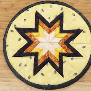 May include: A round quilted potholder featuring a central eight-pointed star pattern. The fabric displays a yellow background with small black bee illustrations, accented by dark brown and orange geometric star points and a solid black border.