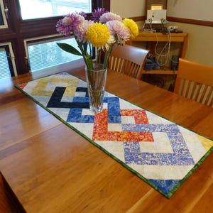 May include: A rectangular quilted table runner featuring a geometric interlocking square pattern in shades of yellow, black, blue, and orange with a white background and a green border, topped with a clear glass vase containing colorful flowers.