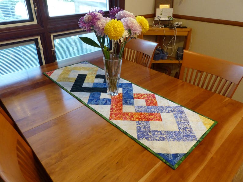 May include: A rectangular quilted table runner featuring a geometric interlocking square pattern in shades of yellow, black, blue, and orange with a white background and a green border, topped with a clear glass vase containing colorful flowers.
