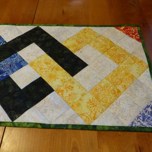 May include: A rectangular quilted table runner featuring a geometric pattern of square fabric patches in shades of blue, black, yellow, and white. The runner is finished with a green fabric border and displays various floral and leaf prints.