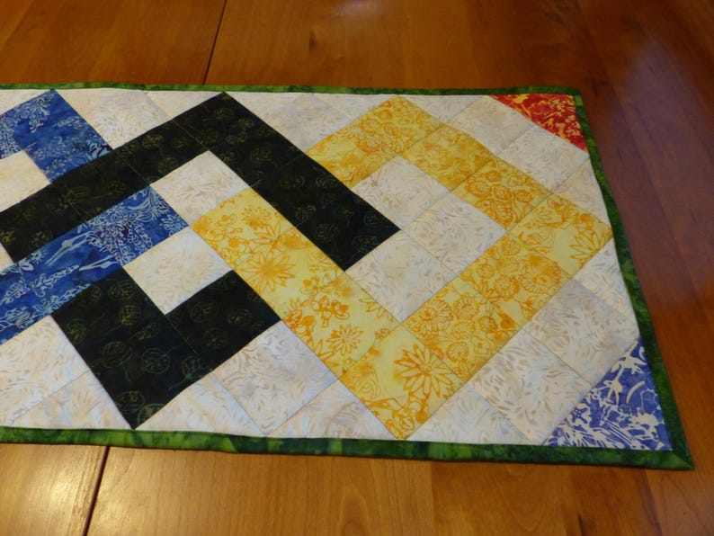 May include: A rectangular quilted table runner featuring a geometric pattern of square fabric patches in shades of blue, black, yellow, and white. The runner is finished with a green fabric border and displays various floral and leaf prints.