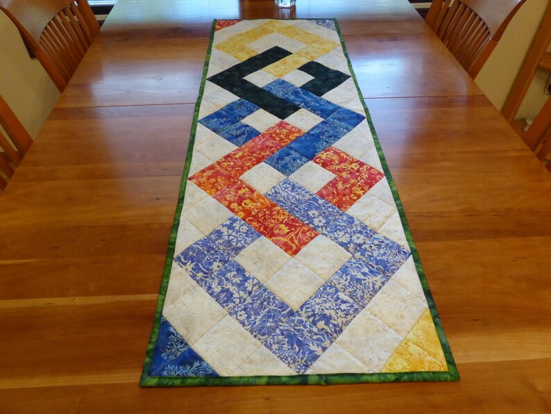 May include: A rectangular quilted table runner featuring a geometric interlocking pattern. The design consists of squares in shades of yellow, black, blue, and red, with floral and mottled fabric textures, finished with a dark green border.