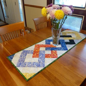 May include: A rectangular quilted table runner featuring a geometric interlocking pattern in shades of blue, orange, yellow, and white, finished with a green border. A clear glass vase containing yellow and purple chrysanthemums sits on top.