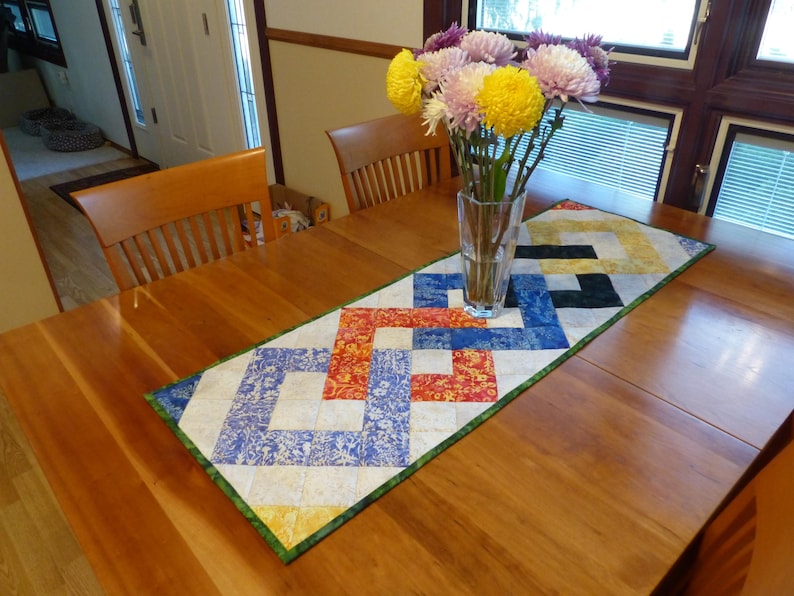 May include: A rectangular quilted table runner featuring a geometric interlocking pattern in shades of blue, orange, yellow, and white, finished with a green border. A clear glass vase containing yellow and purple chrysanthemums sits on top.