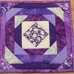 May include: A square, handmade quilted coaster featuring a geometric design in shades of purple, lavender, and white. The center has a floral pattern, and the edges are finished with a coordinating purple binding. The coaster measures approximately 8 inches square.