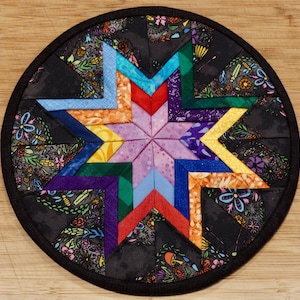 May include: A circular quilted potholder featuring a central star pattern composed of multi-colored fabric segments in shades of purple, blue, green, orange, and red. The outer border consists of black fabric with a colorful floral print.