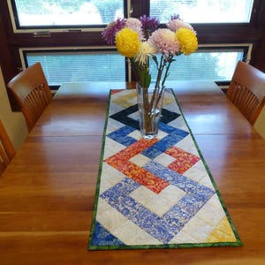 May include: A rectangular quilted table runner featuring a geometric interlocking pattern in shades of blue, red, yellow, and white, with a green border. A clear glass vase containing a bouquet of yellow, white, and purple chrysanthemums sits on top.