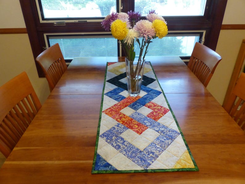 May include: A rectangular quilted table runner featuring a geometric interlocking pattern in shades of blue, red, yellow, and white, with a green border. A clear glass vase containing a bouquet of yellow, white, and purple chrysanthemums sits on top.