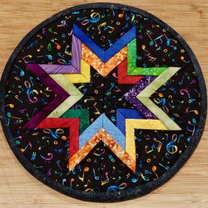 May include: A circular quilted potholder featuring a central eight-pointed star design made of multi-colored fabric segments. The background fabric is black with a colorful pattern of musical notes and symbols. The edge is finished with a dark gray binding.