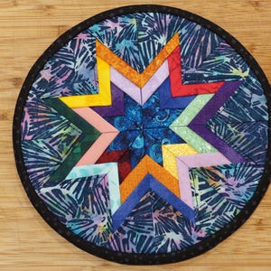 May include: A colorful star-shaped patchwork quilt coaster with a blue and white floral print background. The coaster is round and has a black border.