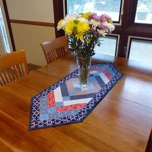 May include: A rectangular quilted table runner featuring a geometric pattern in shades of red, white, and blue with star and floral motifs. A clear glass vase filled with yellow, white, and purple chrysanthemums sits on the center of the runner.