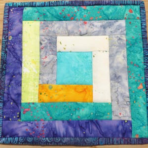May include: Square quilted coaster with a log cabin design. The coaster features a vibrant mix of colors, including teal, purple, yellow, and orange. The edges are finished with a patterned border, and the coaster is placed on a wooden surface.