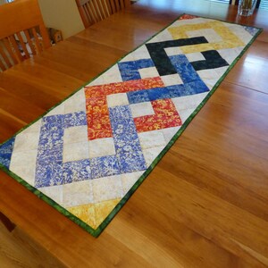May include: A rectangular quilted table runner featuring a geometric interlocking chain pattern. The design incorporates squares of fabric in yellow, black, blue, and red, set against a cream background with a green border trim.