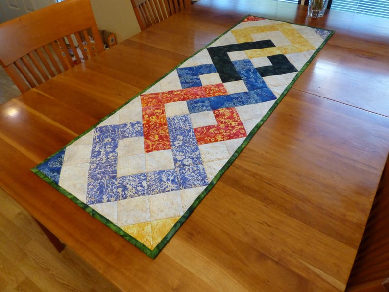May include: A rectangular quilted table runner featuring a geometric interlocking chain pattern. The design incorporates squares of fabric in yellow, black, blue, and red, set against a cream background with a green border trim.
