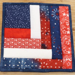 May include: Square quilted coaster with a patriotic design. The coaster features a patchwork pattern with red, white, and blue fabrics. The blue fabrics have a dotted or firework design, while the red fabrics have a star pattern.