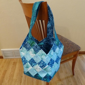 May include: A handmade quilted tote bag in shades of blue and teal. The bag features a patchwork design with a diamond pattern. The handles are a solid teal color. The bag is hanging on a wooden chair.