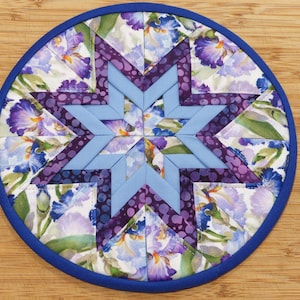 May include: Circular quilted trivet with a blue border. The center features a light blue star design surrounded by purple dotted fabric and floral print fabric with purple and blue irises. A decorative kitchen item.