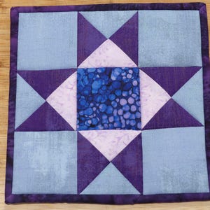 May include: A square fabric quilt block featuring a central square with a blue and purple bubble pattern, surrounded by light purple triangles and light blue squares. The entire piece is framed by a dark purple fabric border with stitched edges.