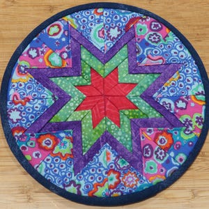 May include: A round, quilted hot pad with a star design. The star is composed of red, green, and purple fabric pieces. The background is a mix of blue, pink, and green floral patterns, with a dark blue border.