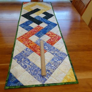May include: A rectangular quilted table runner featuring a geometric pattern of interlocking squares in yellow, black, blue, and red floral fabrics, bordered in green. A wooden ruler is placed on the runner, showing a length of 12 inches.