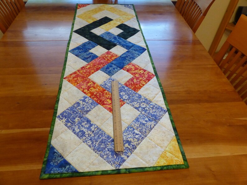 May include: A rectangular quilted table runner featuring a geometric pattern of interlocking squares in yellow, black, blue, and red floral fabrics, bordered in green. A wooden ruler is placed on the runner, showing a length of 12 inches.