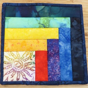 May include: A square, handmade fabric coaster with a patchwork design. The coaster features a vibrant mix of colors, including blue, green, yellow, orange, red, and white. The edges are finished with a dark blue binding. The design includes a sunburst pattern.