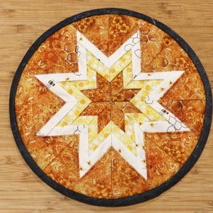May include: A round, quilted coaster with a star design. The star is made of white and yellow fabric, and the background is a patterned orange fabric. The coaster has a black border and is placed on a wooden surface.