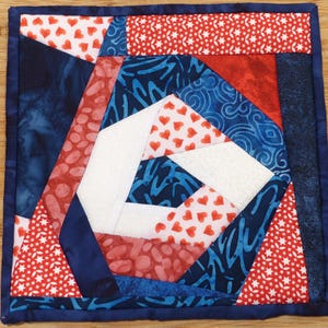 May include: A square, handmade quilted coaster with a patriotic theme. The design features red, white, and blue fabrics in geometric shapes, including hearts and stars. The coaster is bordered with a dark blue fabric.