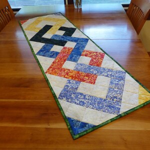 May include: A rectangular quilted table runner featuring a geometric interlocking pattern. The design incorporates fabric squares in shades of yellow, black, blue, orange, and white with floral motifs, all bordered by a green fabric trim.