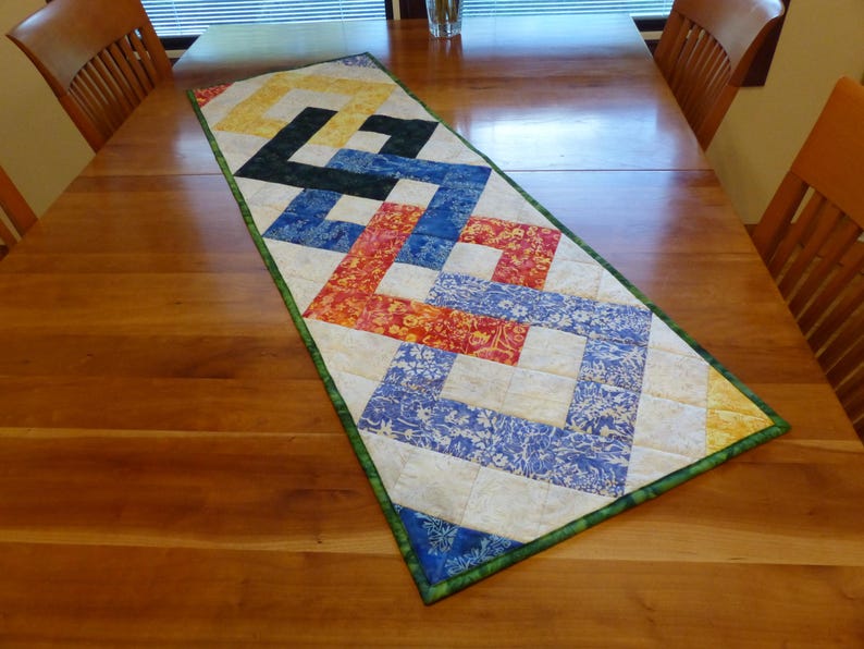 May include: A rectangular quilted table runner featuring a geometric interlocking pattern. The design incorporates fabric squares in shades of yellow, black, blue, orange, and white with floral motifs, all bordered by a green fabric trim.