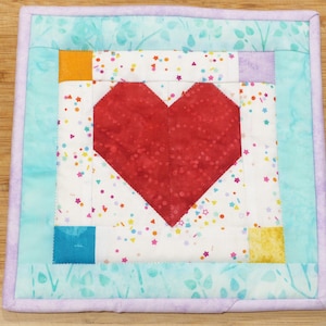 May include: A handmade quilted heart-shaped coaster with a red heart in the center, surrounded by a white background with colorful confetti. The coaster is framed by a turquoise border and a lavender outer border.