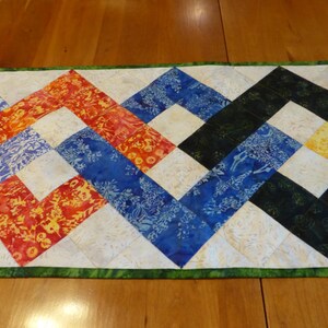 May include: A rectangular quilted table runner featuring a vibrant zigzag pattern composed of multi-colored fabric squares. The design includes shades of orange, red, blue, black, and yellow with floral motifs, separated by cream-colored squares and a green border.