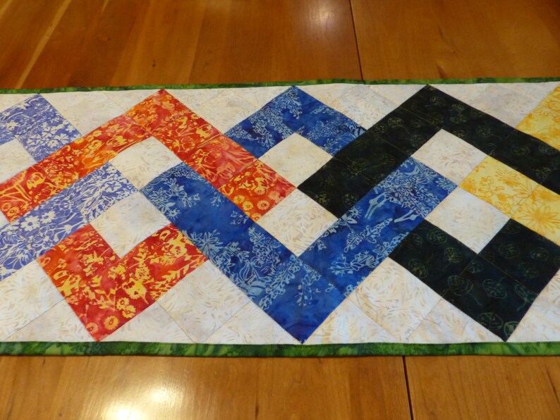 May include: A rectangular quilted table runner featuring a vibrant zigzag pattern composed of multi-colored fabric squares. The design includes shades of orange, red, blue, black, and yellow with floral motifs, separated by cream-colored squares and a green border.