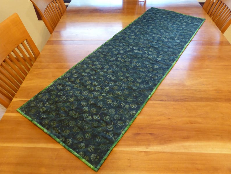 May include: A rectangular quilted table runner featuring a dark green fabric base with a repeating pattern of light green leaves. The runner has a thin, bright green fabric border along all four edges and a visible quilted texture throughout.