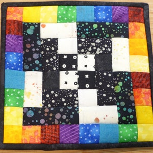 May include: A colorful patchwork quilt square with a black and white checkered pattern in the center. The square is surrounded by a border of colorful fabric strips in shades of red, orange, yellow, green, blue, and purple.