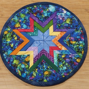 May include: Circular, quilted trivet featuring a vibrant star design. The star is composed of various colorful fabrics, including blue, orange, yellow, green, and purple. The background is a blue fabric with a speckled pattern, and the edge is a dark blue binding.