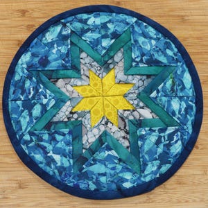 May include: Circular quilted trivet with a star design. The trivet features a yellow star in the center, surrounded by teal and blue geometric shapes. The outer edge is a dark blue, and the background is a light wood grain.