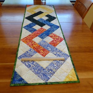 May include: A rectangular quilted table runner featuring a geometric interlocking pattern in yellow, black, blue, and red floral fabrics on a cream background, with a green border and a clear ruler showing a width of approximately 12 inches.