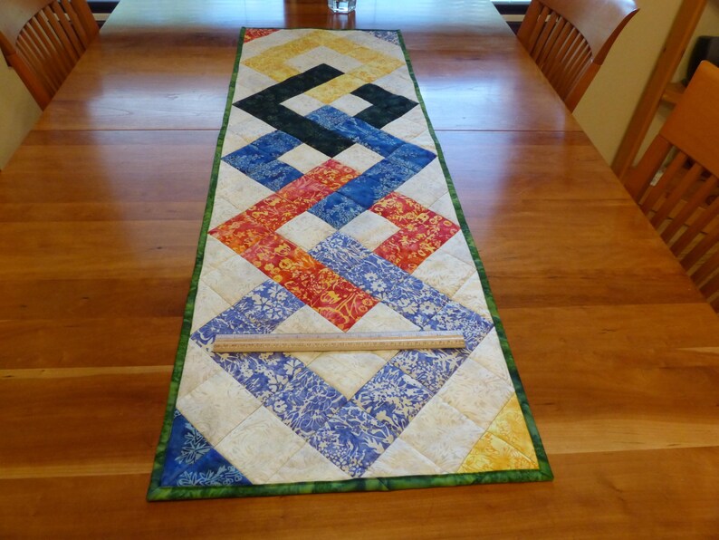 May include: A rectangular quilted table runner featuring a geometric interlocking pattern in yellow, black, blue, and red floral fabrics on a cream background, with a green border and a clear ruler showing a width of approximately 12 inches.