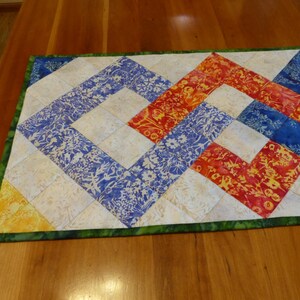 May include: A rectangular quilted table runner featuring a geometric pattern of interconnected squares. The design incorporates batik-style fabrics in vibrant shades of blue, orange, yellow, and cream, finished with a green patterned fabric border.