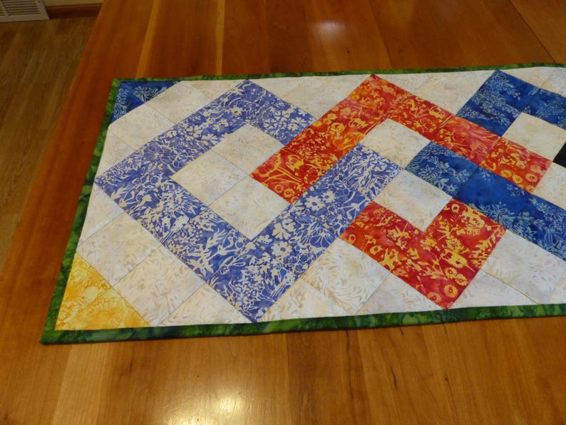 May include: A rectangular quilted table runner featuring a geometric pattern of interconnected squares. The design incorporates batik-style fabrics in vibrant shades of blue, orange, yellow, and cream, finished with a green patterned fabric border.