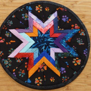 May include: A round, quilted coaster with a black background featuring colorful paw prints. The center has a star design made of various fabric pieces in shades of red, orange, blue, purple, and pink. The coaster is approximately 5 inches in diameter.