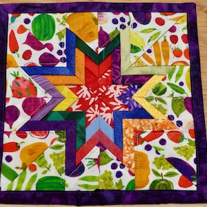 May include: A square fabric potholder featuring a central star pattern constructed from multi-colored fabric strips. The background fabric displays a vibrant print of various fruits and vegetables, including watermelons, carrots, and grapes. The entire piece is finished with a solid purple fabric border.