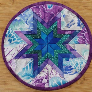May include: A round, quilted trivet with a starburst design. The star is composed of various shades of blue, teal, and purple fabrics. The trivet is edged with a purple binding. The background is a light brown wooden surface.