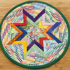 May include: A round, green-trimmed, quilted hot pad with a colorful star design. The star is made of fabric squares in various shades of blue, purple, green, yellow, orange, and red. The background fabric is white with a rainbow polka dot pattern.