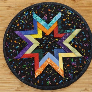 May include: A round, quilted trivet with a black background covered in colorful musical notes. A vibrant star design in shades of blue, orange, yellow, green, purple, and red is centered on the trivet. The trivet has a dark gray border.