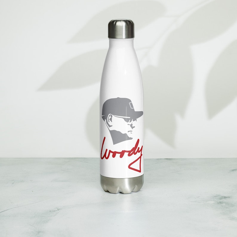 Puede incluir: Botella de agua de acero inoxidable blanca con una silueta gris de una persona con sombrero y gafas de sol. La palabra "Woody" est&aacute; escrita en cursiva roja debajo de la silueta.