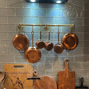 Peut inclure: Une collection d'ustensiles de cuisine en cuivre comprenant une barre de suspension avec six casseroles, une bouilloire et une petite marmite avec couvercle. Une planche à découper en bambou avec le texte KITCHEN WITCHERY et des cuillères en bois.