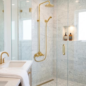 Puede incluir: Un baño con ducha a ras de suelo con paredes de azulejos de mármol blanco y suelo de mármol hexagonal. La ducha incluye un cabezal de ducha tipo lluvia dorado, un rociador manual y una puerta de cristal. Un grifo dorado sobre un tocador blanco.